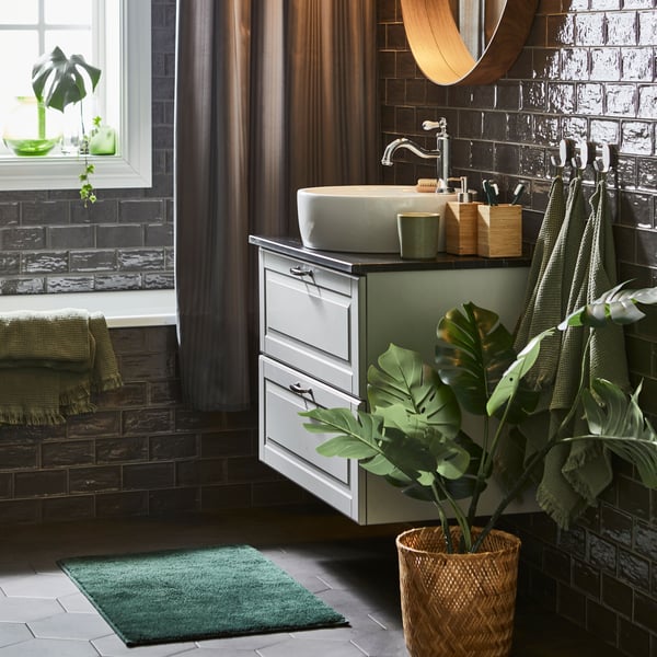 A dark green SÖDERSJÖN bath mat complements a stylish bathroom featuring dark tiles, a circular mirror, and plants.