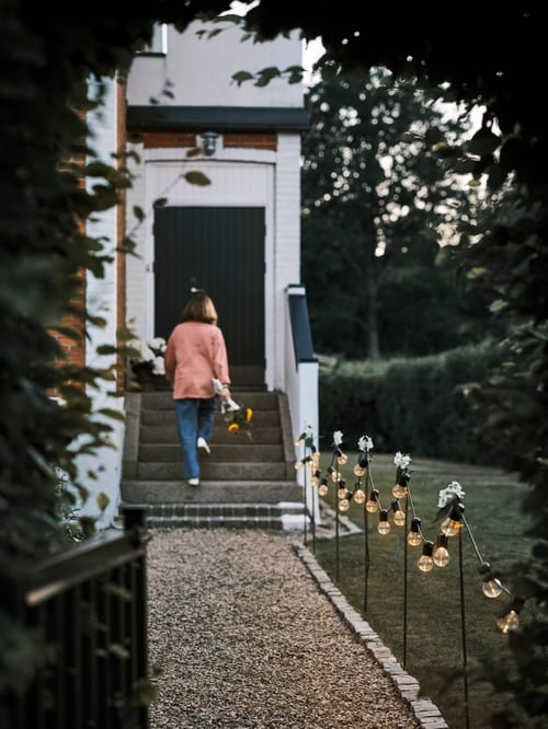 A chain of SVARTRÅ lighting hangs from poles along a gravel path, and a person holding flowers walks up stairs toward a door.