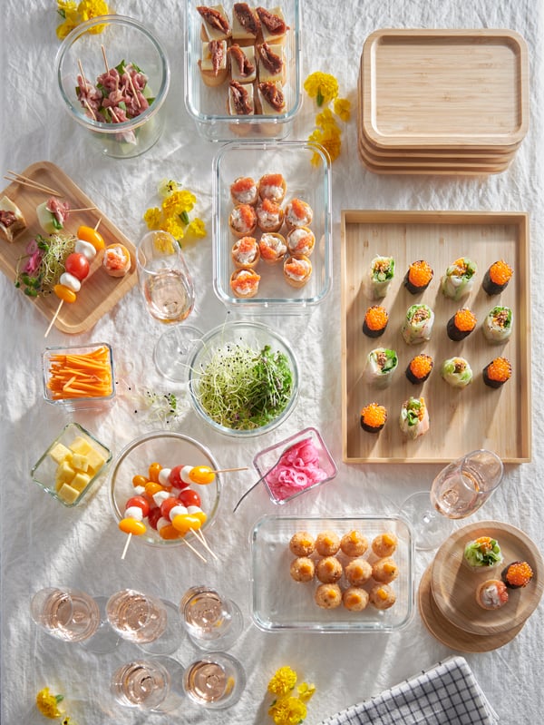 Una tavola con una tovaglia bianca allestita con ciotole, contenitori IKEA 365+ e vassoi OSTBIT con tantissimi alimenti come sushi vegetariano, radici, verdure e altro. Ci sono anche sottobicchieri e coperchi IKEA 365+ impilati l'uno sull'altro.