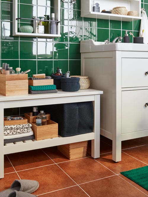 Modern bathroom featuring a white bench, a wall shelf, several baskets, boxes with lids, a wash stand and towels. 