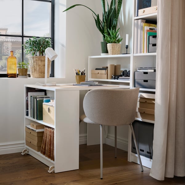 Corner of a bright room with BILLY bookcases, a section on one of which has been opened to create a desk that is full of desk accessories.