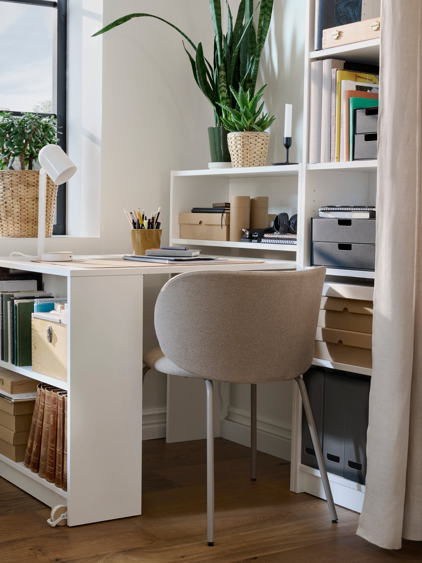 A corner of a bright room with BILLY bookcases, with one section is pulled out to form a desk, filled with desk accessories.