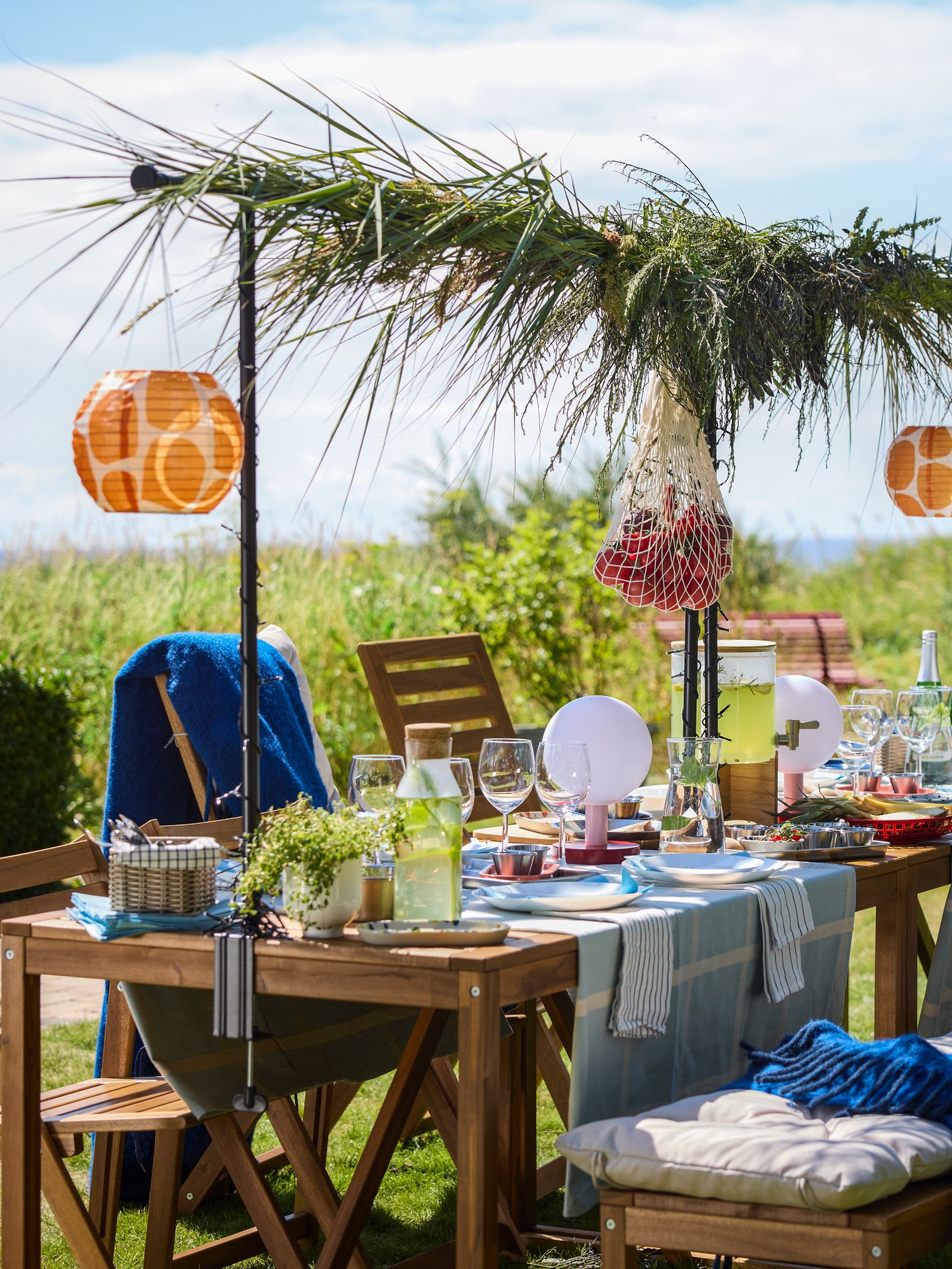 Słoneczny dzień z ogrodem z jasnobrązowym drewnianym stołem NÄMMARÖ, na którym stoją potrawy i napoje. Krzesła, ławka i parasol.