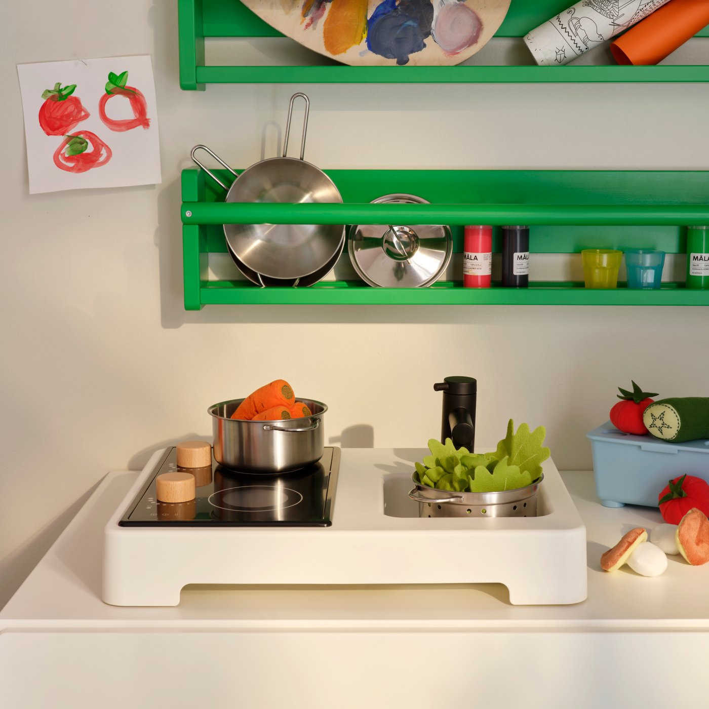 Parts of a STENMUS mini-play kitchen beneath green FLISAT wall storage, all filled with play-cooking accessories.