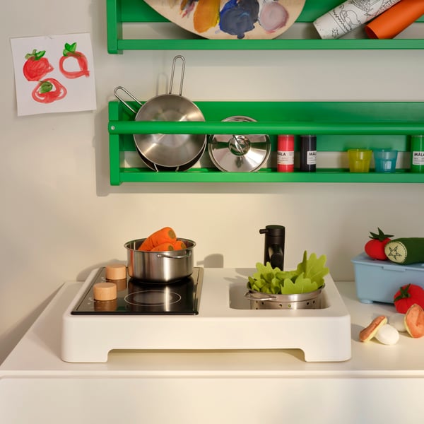 Parts of a STENMUS mini-play kitchen beneath green FLISAT wall storage, all filled with play-cooking accessories.