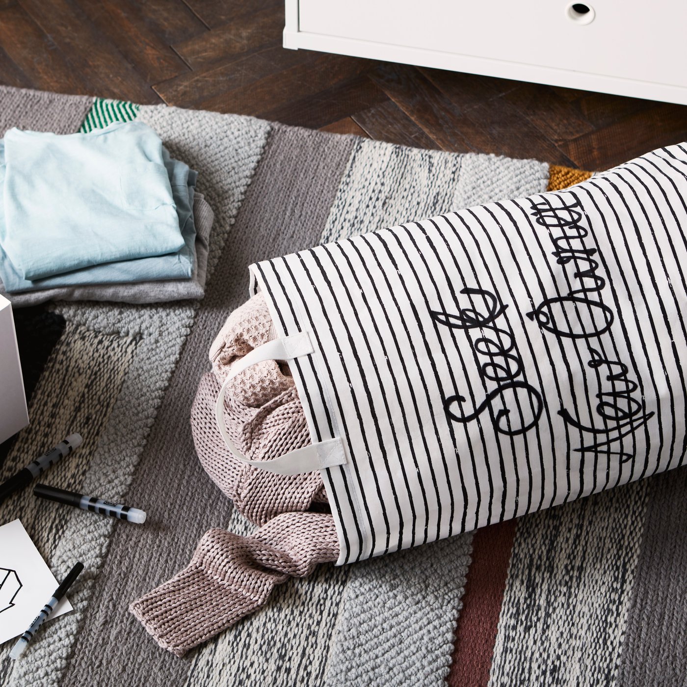 Ein KLUNKA Wäschesack gefüllt mit Pullovern auf einem grauen Teppich. Daneben sind ein weisser Kasten und weitere Kleidungsstücke zu sehen.