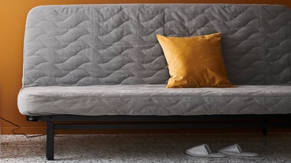 Grå NYHAMN bäddsoffa med quiltad klädsel och dekorativ kudde framför färgad vägg.