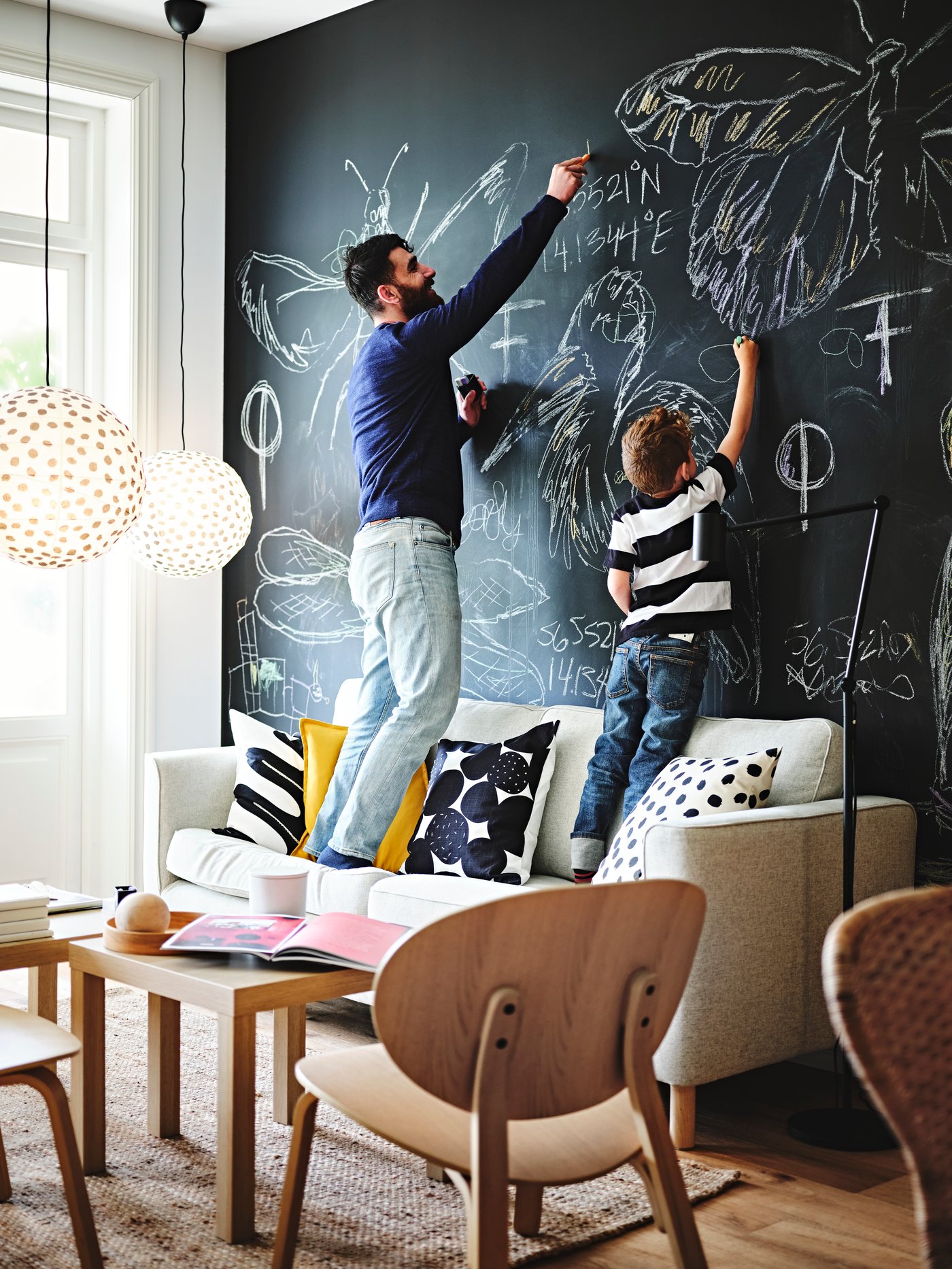 Un enfant et un adulte sont debout sur un canapé pour dessiner des images sur un tableau noir. Deux abat-jour REGOLIT se trouvent derrière eux.