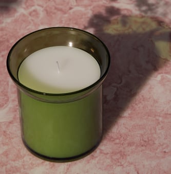 HEDERSAM mirisna sveća u zelenom staklenom držaču postavljenom na roze površinu.