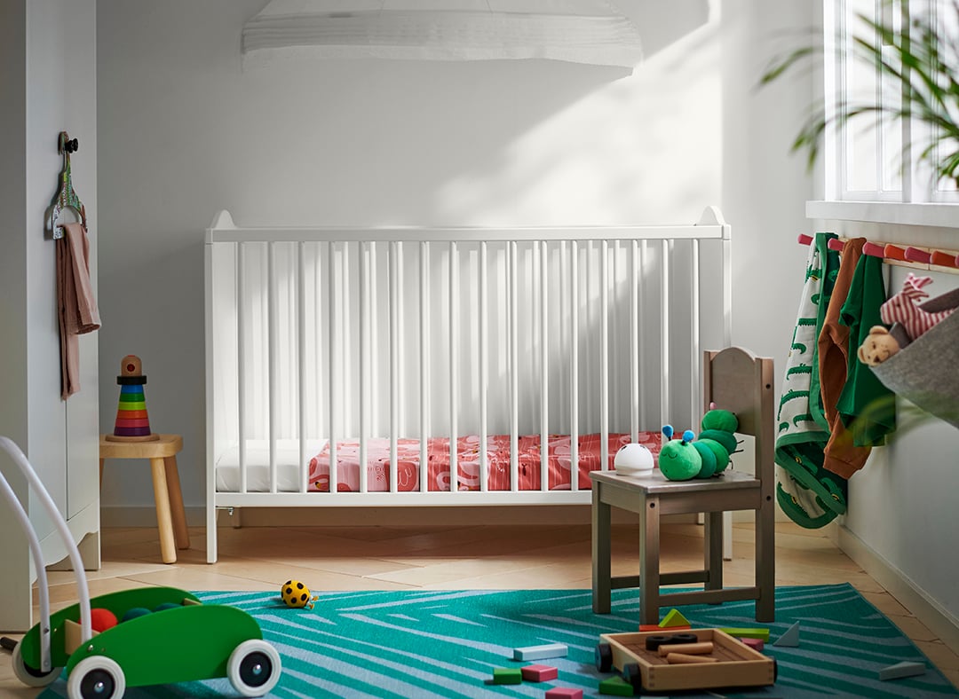 Un lettino SMÅGÖRA bianco con sopra un baldacchino posto in una cameretta per neonato con davanti un tappeto azzurro e alcuni giocattoli in legno.