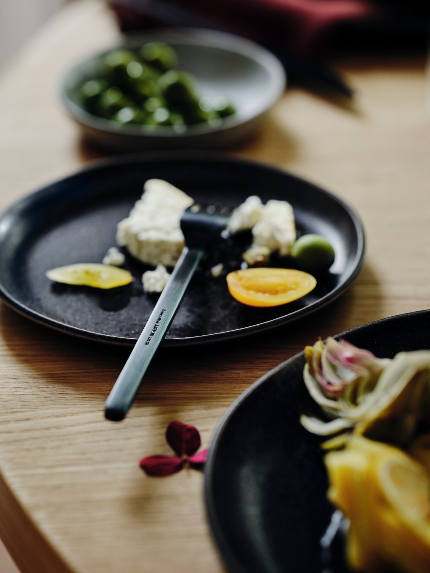 Dubová deska stolu s černým talířem STOCKHOLM 2025, malým talířkem a servírovacím talířem. Talíř obsahuje jídlo a příbory.