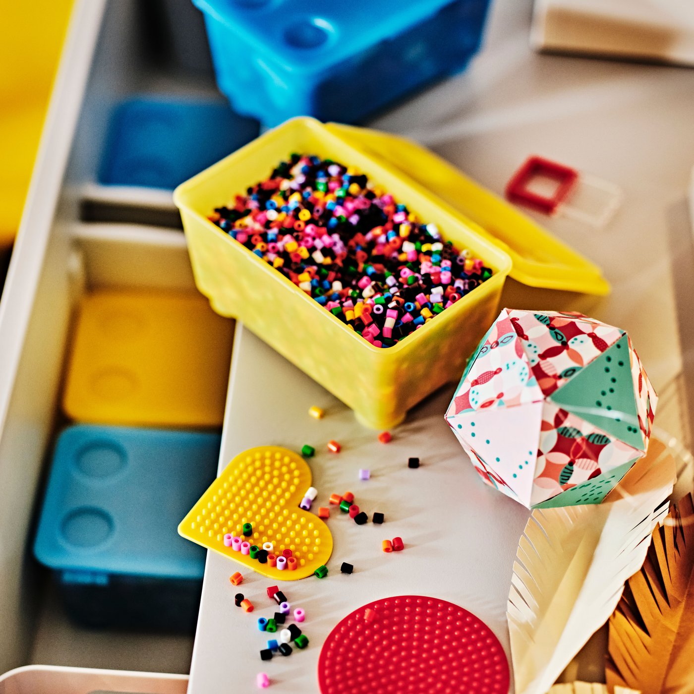 Bunte Perlen in gelbem Behälter mit geometrischen Formen wie Herz und Polyeder, daneben blaue und gelbe Aufbewahrungsboxen.