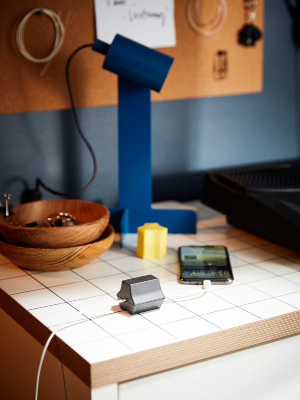 A MICKE desktop with a dark-blue FLOTTILJ lamp and a charging phone, the cord held in place with a HAVSKÅL USB anchor.