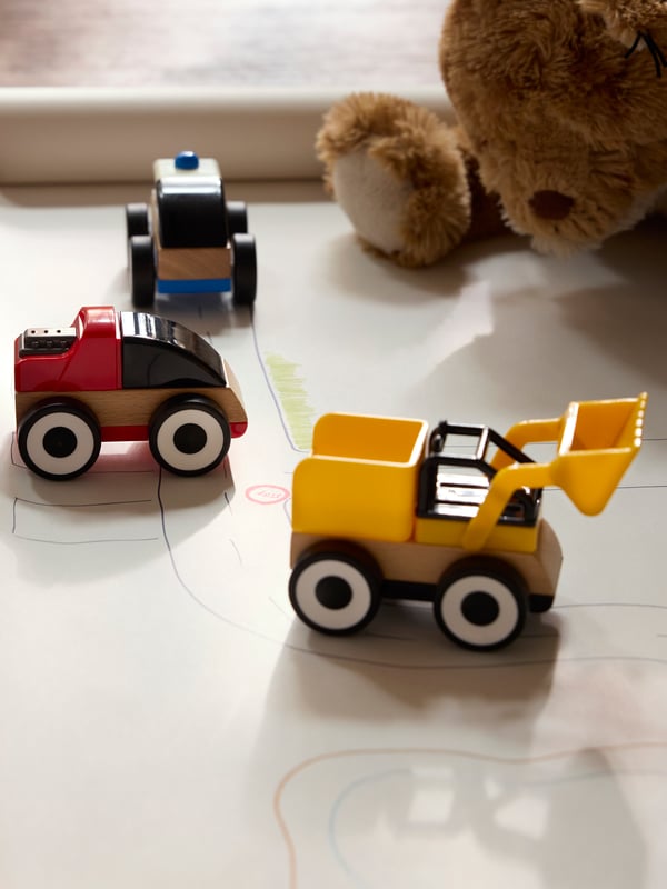 Close-up of three LILLABO toy vehicles in yellow, red and blue. Teddy bear sitting behind.