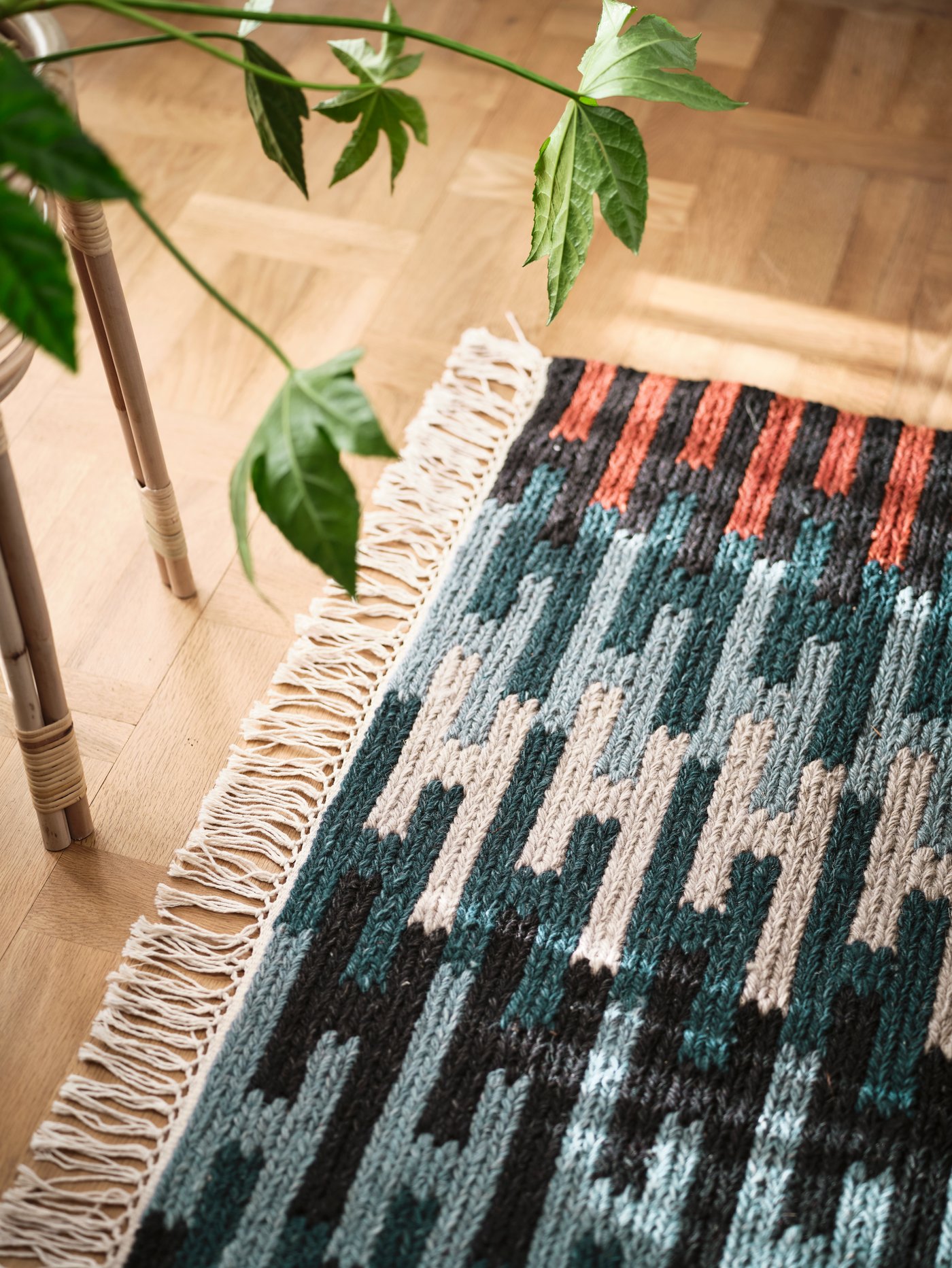 A light wooden floor with a flatwoven RESENSTAD rug in different shades of blue, grey and red and a plant in a plant stand.