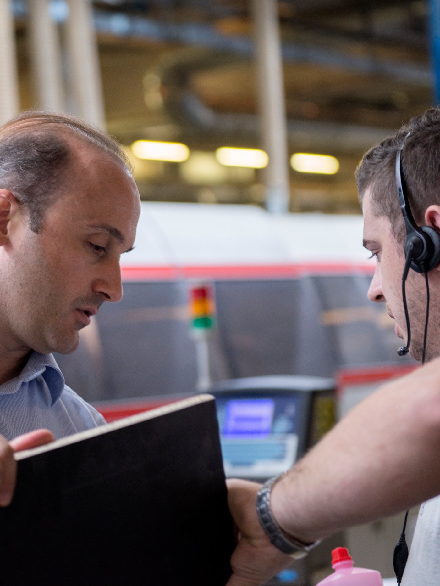 Two men standing beside some machinery in a factory, one carrying a lap top computer and the other wearing a headset.