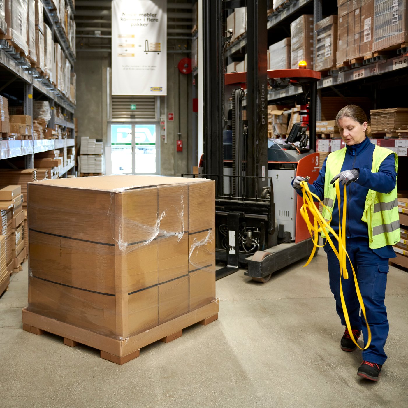 Person med varselväst går genom ett lagerutrymme och håller i gula spännband. I mitten står en stor plastinplastad pall med kartonger. Hyllrader fyllda med lådor syns på båda sidor och en truck står bakom pallen.