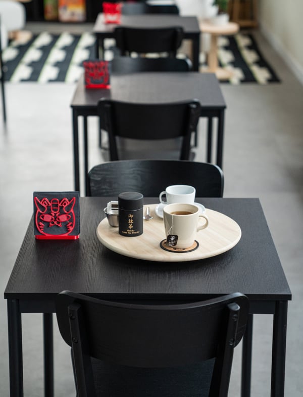 Una fila di tre tavoli e sedie nere. Su quello in primo piano c'è un vassoio con con sopra due tazze una di caffè e una di tè.