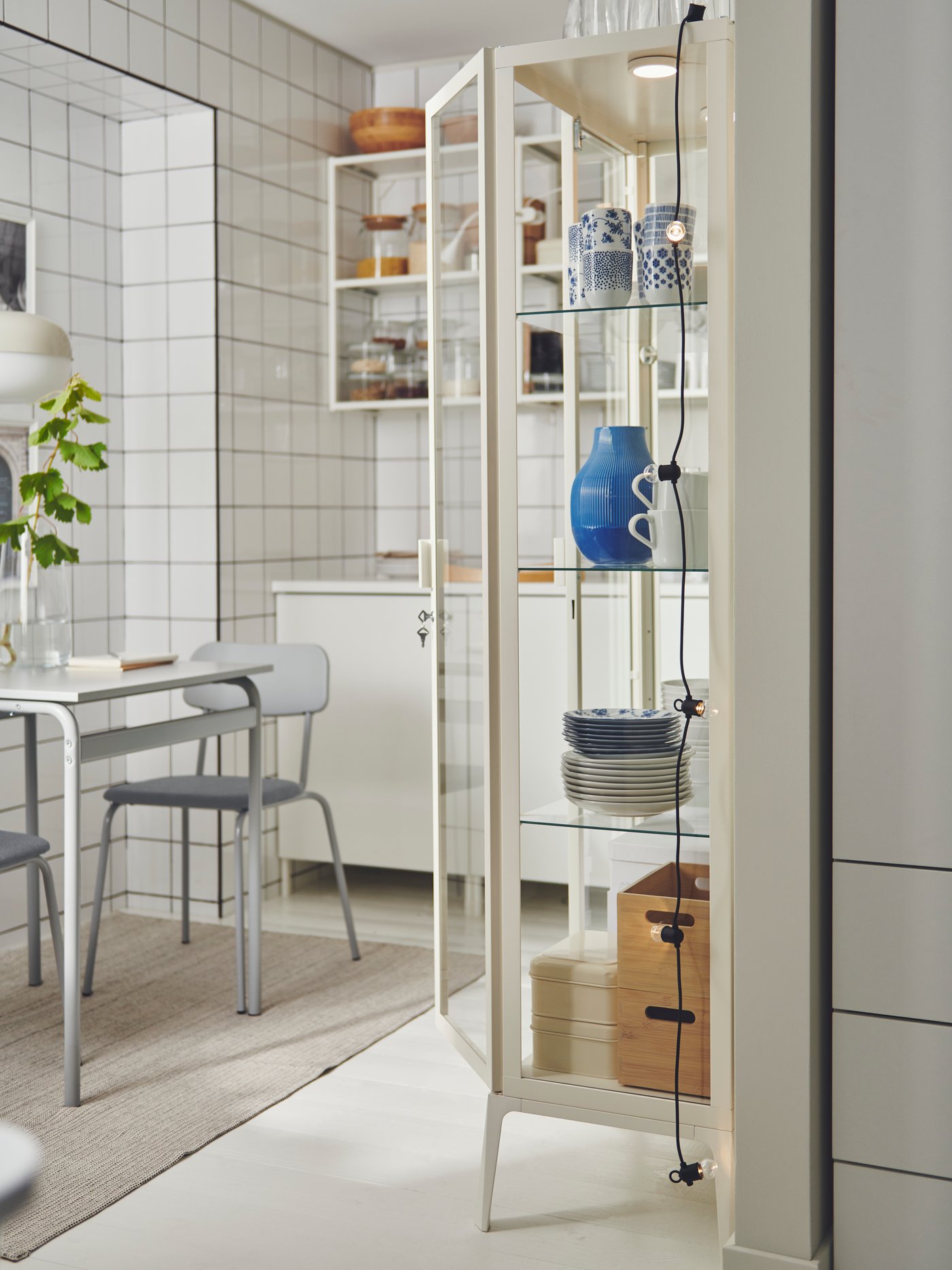 Una cocina con una vitrina MILSBO con un jarrón azul y tazas ENTUSIASM. A su lado hay una mesa con dos sillas.