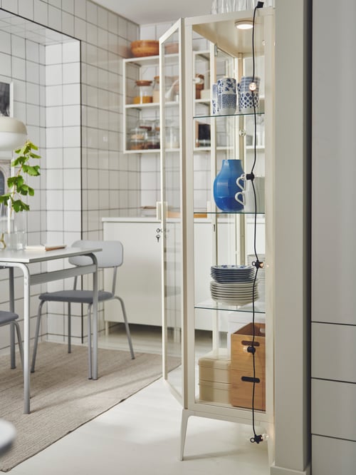 A kitchen with a MILSBO glass door cabinet filled with a blue vase and ENTUSIASM mugs. Beside it, a table with two chairs.