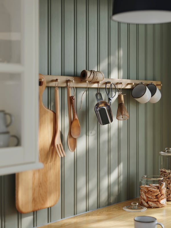 A green kitchen wall with HÖVOLM racks with 6 knobs in oak on the wall, GLADELIG mugs in grey and items hanging from them.