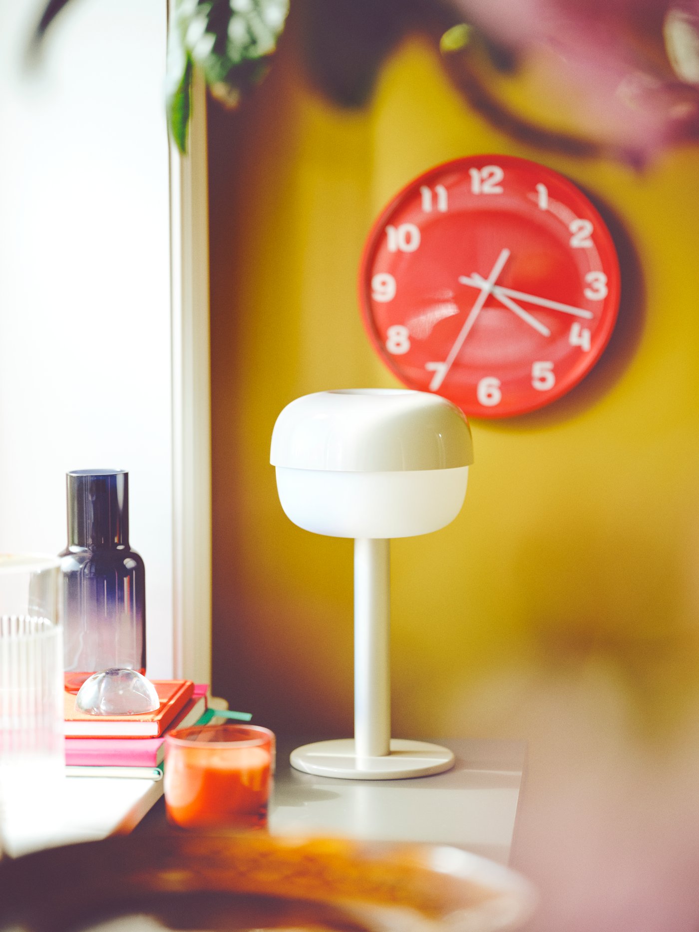 Una lámpara de mesa BLÅSVERK blanca sobre una mesa; junto a ella, hay un jarrón FÖRENLIG gris. Detrás, hay un reloj de pared PLUTTIS rojo.