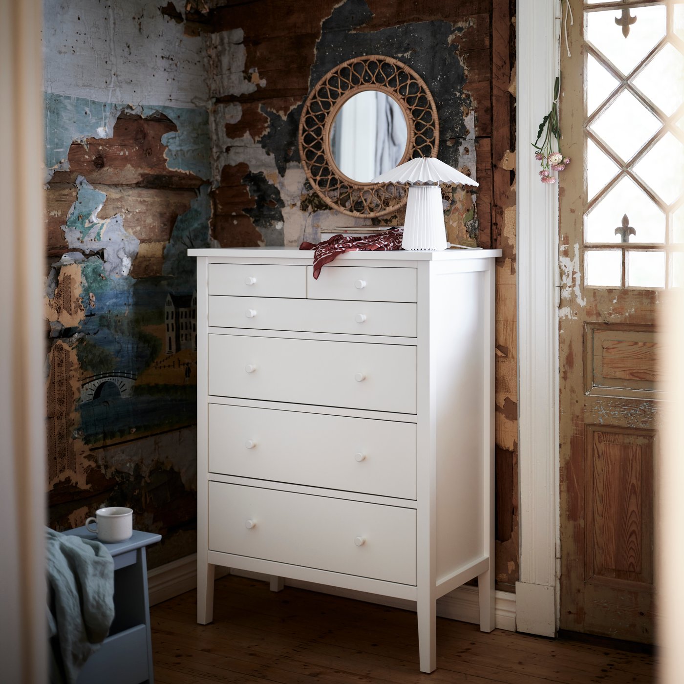 En hvit GULLABERG-kommode med 6 skuffer står mot en rustikk vegg med et rundt speil, lampe og småting arrangert på toppen.