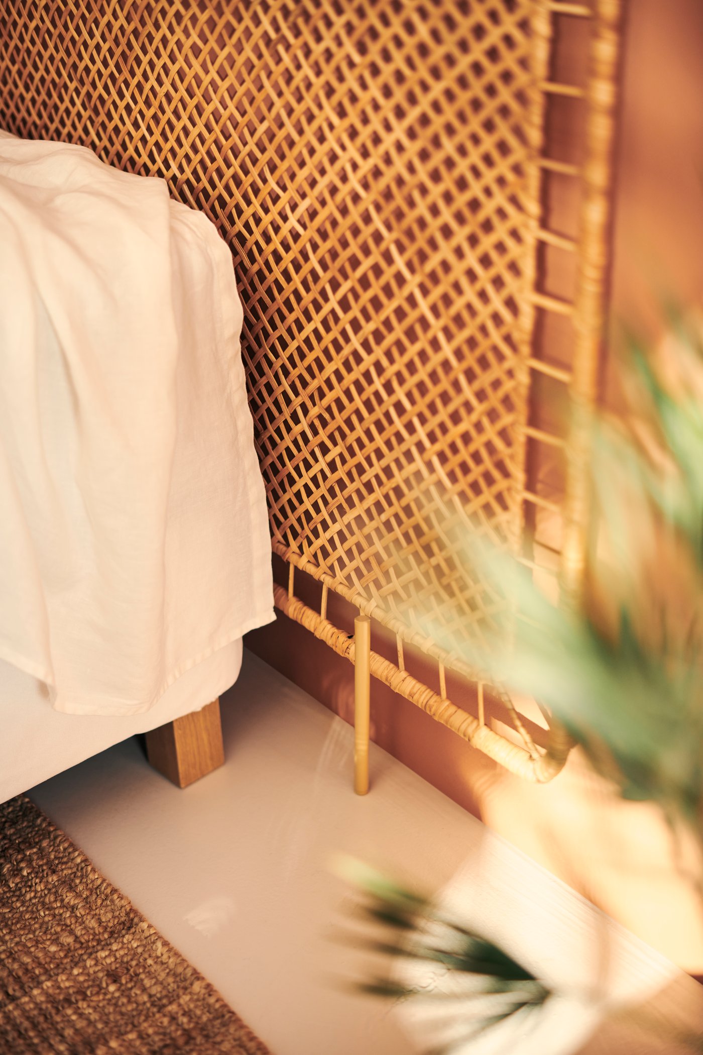 En närbild av en sänggavel i flätad rotting bredvid en säng med vit textil, med varmt ljus och suddiga gröna växter i förgrunden.
