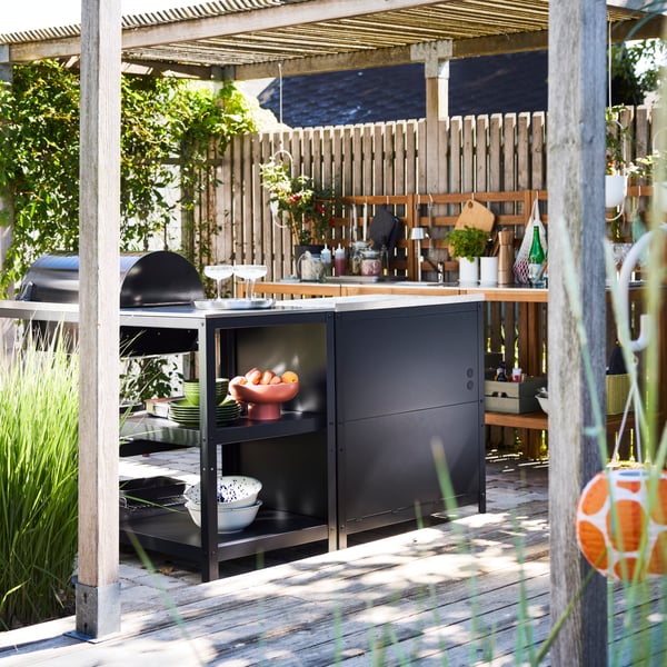 A large wooden patio with plants and lamps and a black GRILLSKÄR charcoal barbecue, part of a kitchen island setup.