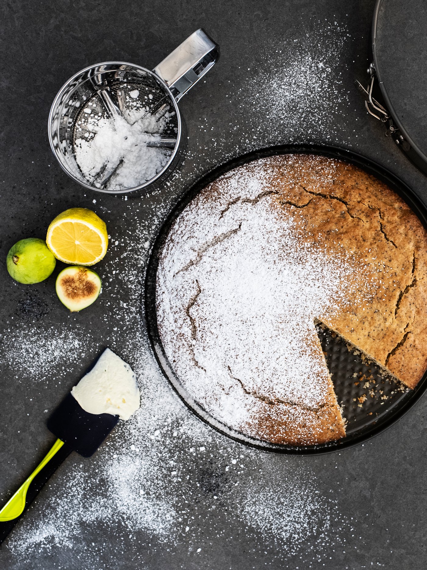 Vista superior de un molde HEMMABAK con un queque preparado, una espátula de goma GUBBRÖRA y un cernidor IDEALISK metálico con azúcar flor, dispuestos sobre una cubierta de cocina gris oscura. 