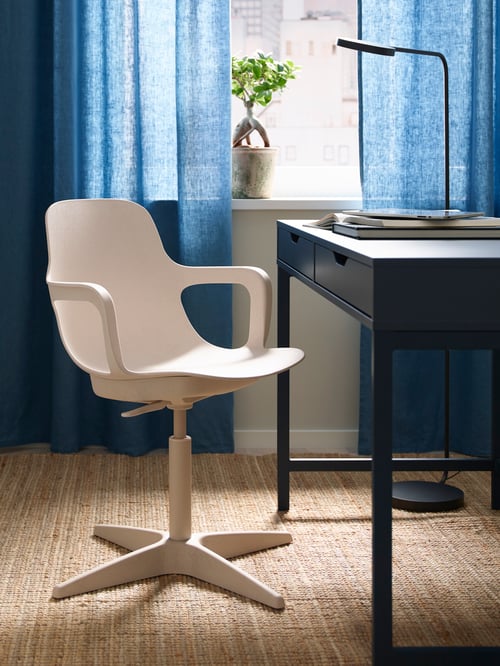 A white/beige ODGER swivel chair is on a jute rug by a blue desk, in front of a curtained window. The desk has books on it.