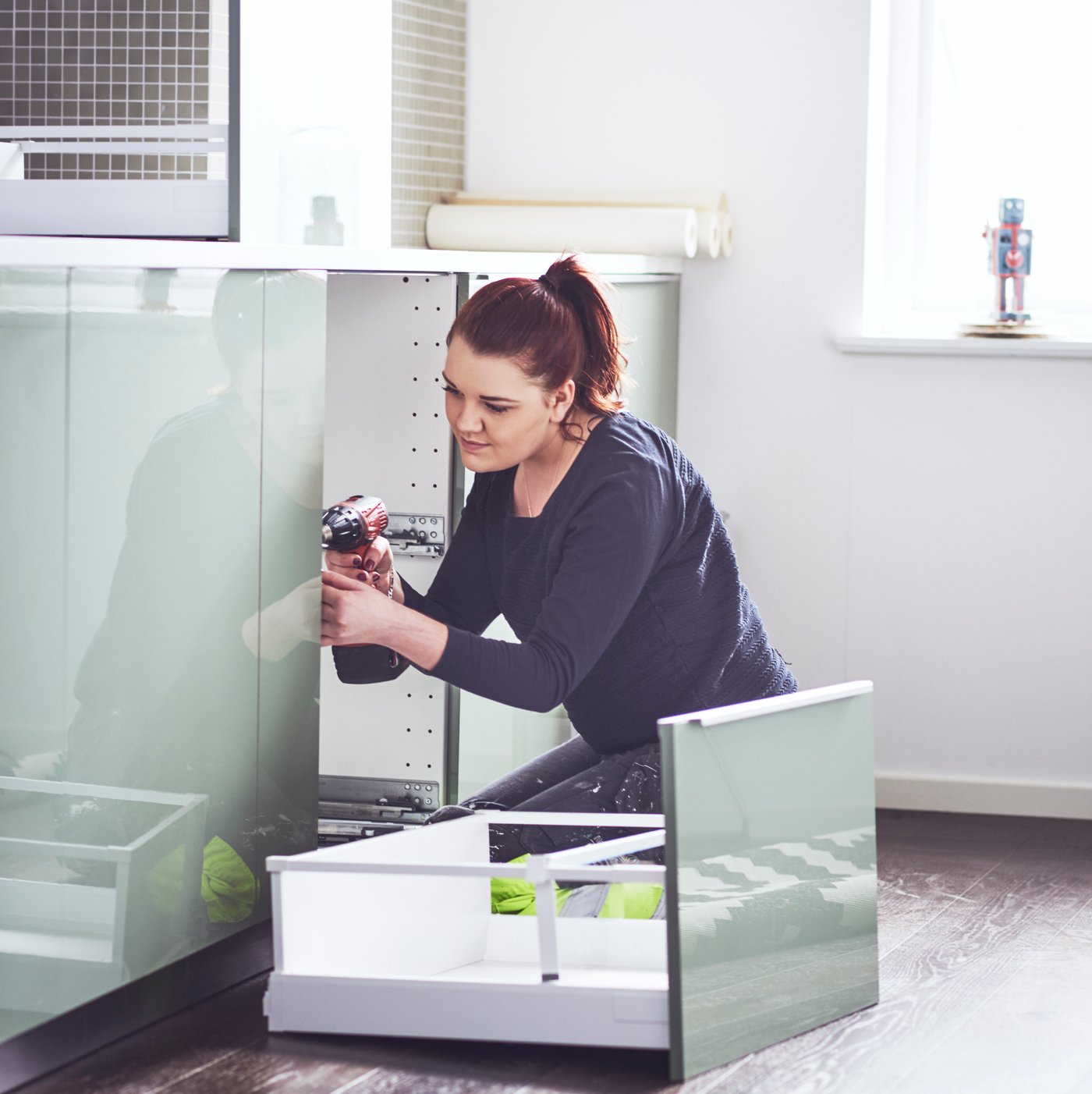 Woman with electric drill kneeling down and assembling a kitchen drawer
