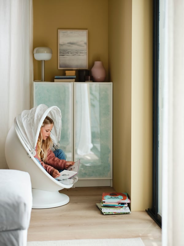 A child is sitting and reading inside an IKEA PS LÖMSK swivel armchair next to a bookcase combination with glass doors.