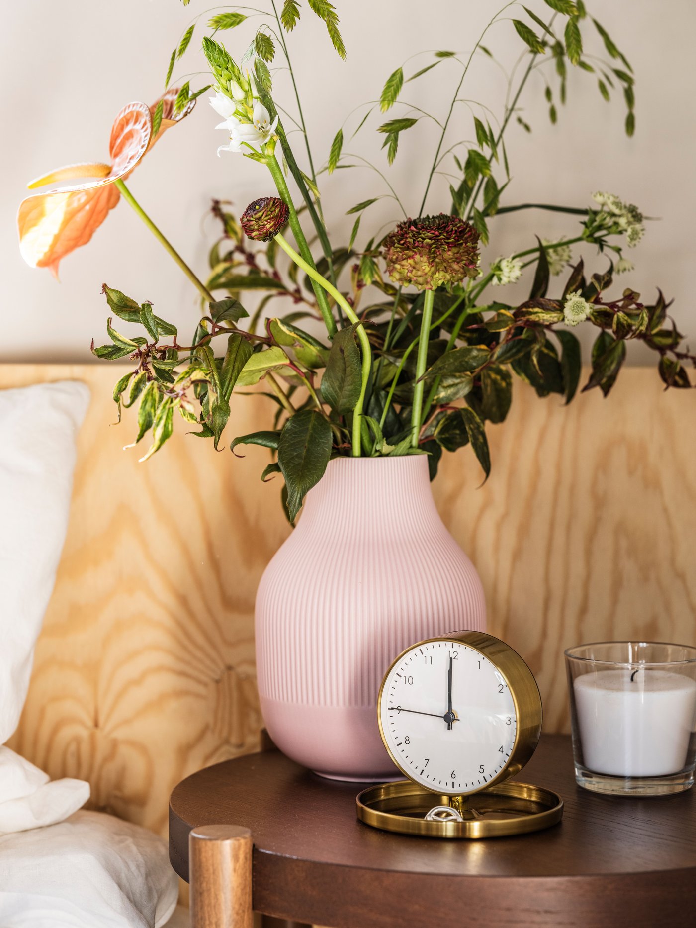 Een roze GRADVIS vaas naast een goudkleurige wekker op een bijzettafel naast een bed in de slaapkamer.