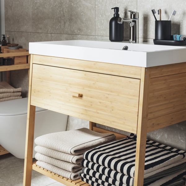Ett RÅGRUND tvättställ i naturfärgad bambu med en låda och ett öppet hyllplan. Beige och randiga handdukar vikta i två högar.