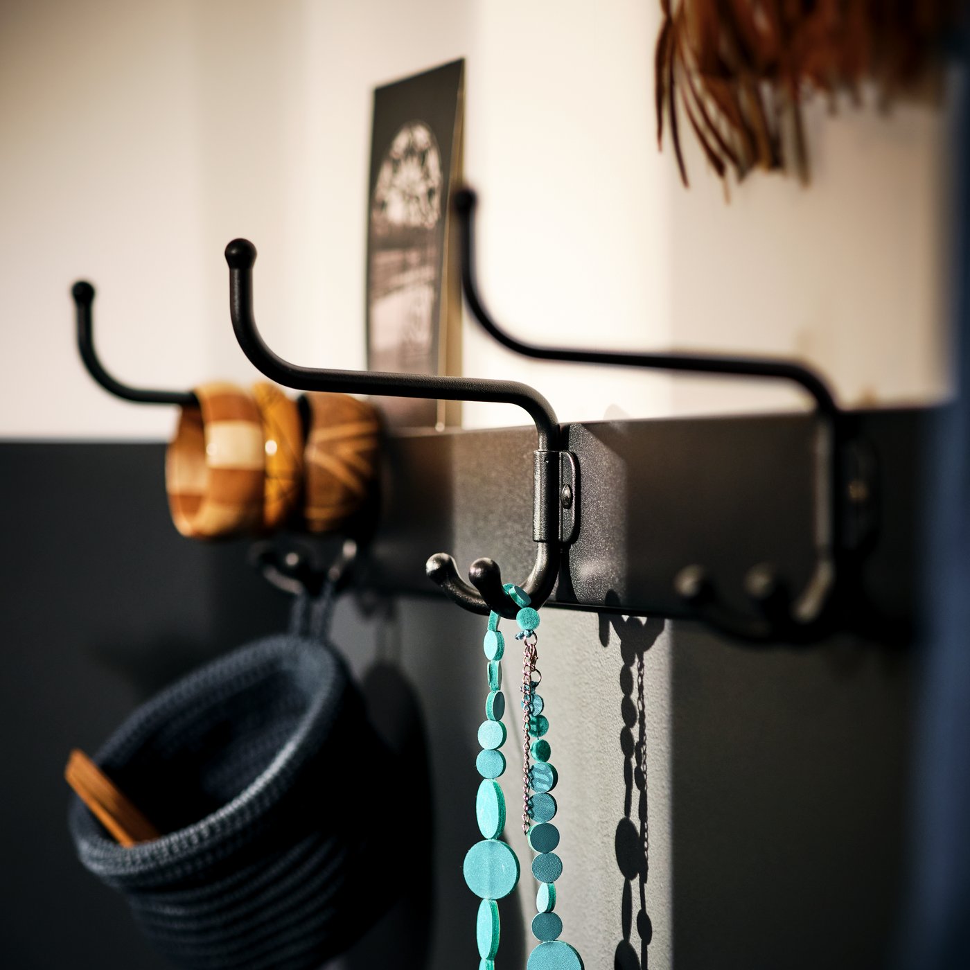 En rad svarta metallkrokar på väggen, dekorerade med små föremål som armband, halsband och en stickad mössa, i varmt infallande ljus.