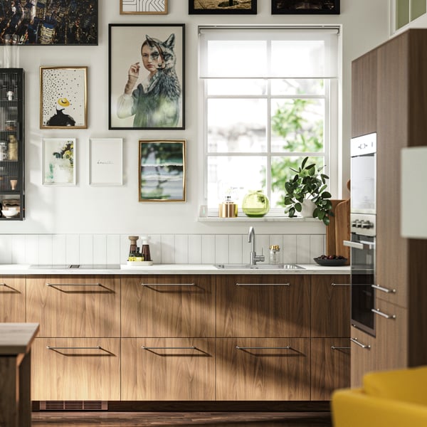 A kitchen with TISTORP fronts with a brown walnut effect, a dark wood floor and some posters on the wall next to a window