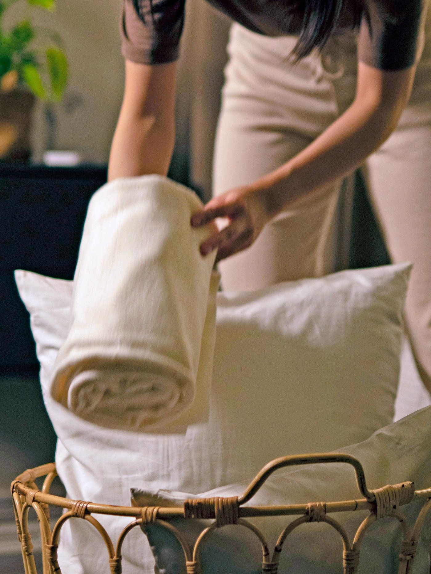 Una mujer coloca un cojín KÄRLEKSGRÄS, una manta THORGUN y una almohada LAPPTÅTEL en una cesta que está en el suelo junto a la cama.