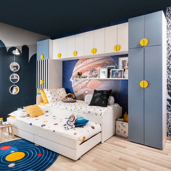 Una camera da letto con pareti decorate con diverse illustrazioni sulle pareti. Al centro una struttura armadio attorno a due letti con lenzuola bianche. Accanto al letto uno sgabellino FLISAT e un contenitore  con alcuni elementi al suo interno.