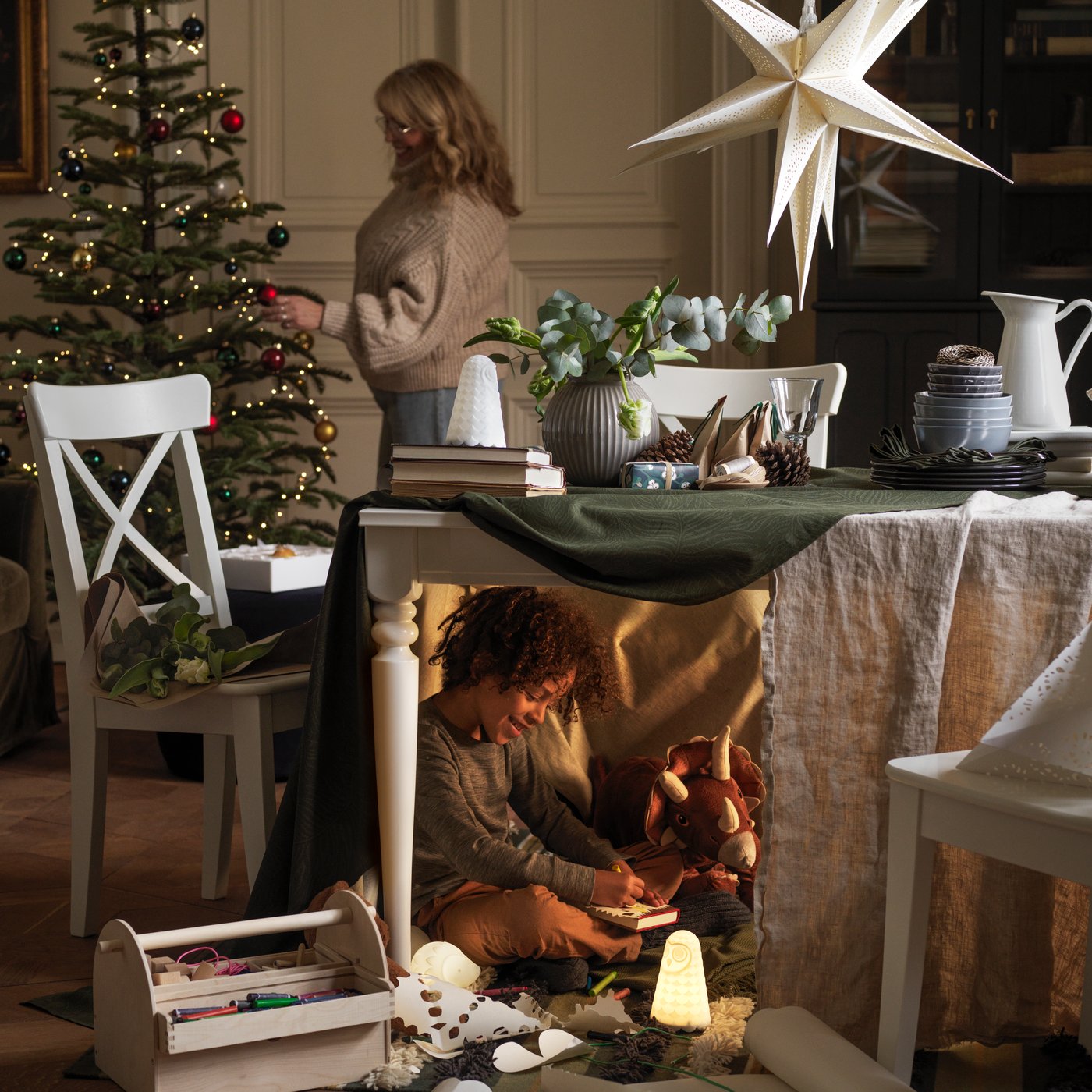 Una persona sta decorando l'albero di Natale e un bambino sta giocando con dei giocattoli seminascosti sotto il tavolo in primo piano.