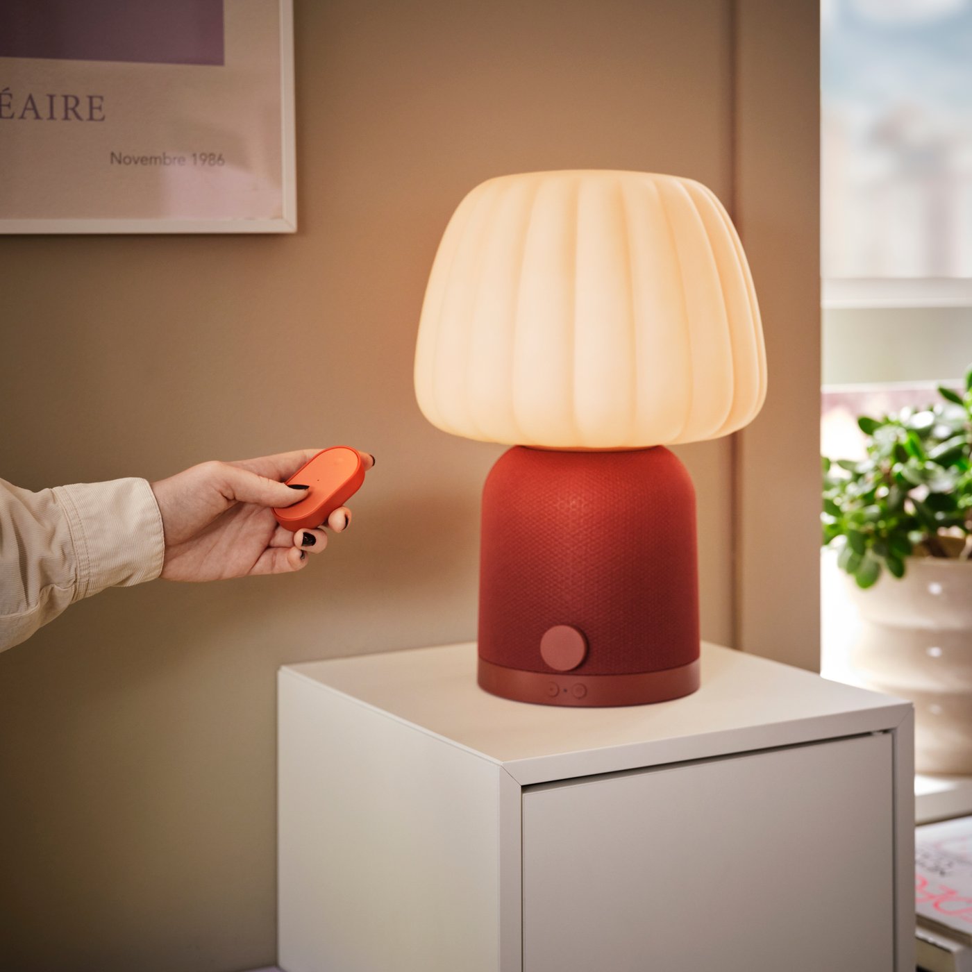 En bordslampa med rundad, veckad lampskärm som lyser varmt ovanpå en röd bas. Lampan står på ett vitt skåp mot en beige vägg. En hand håller en liten röd fjärrkontroll nära lampan.