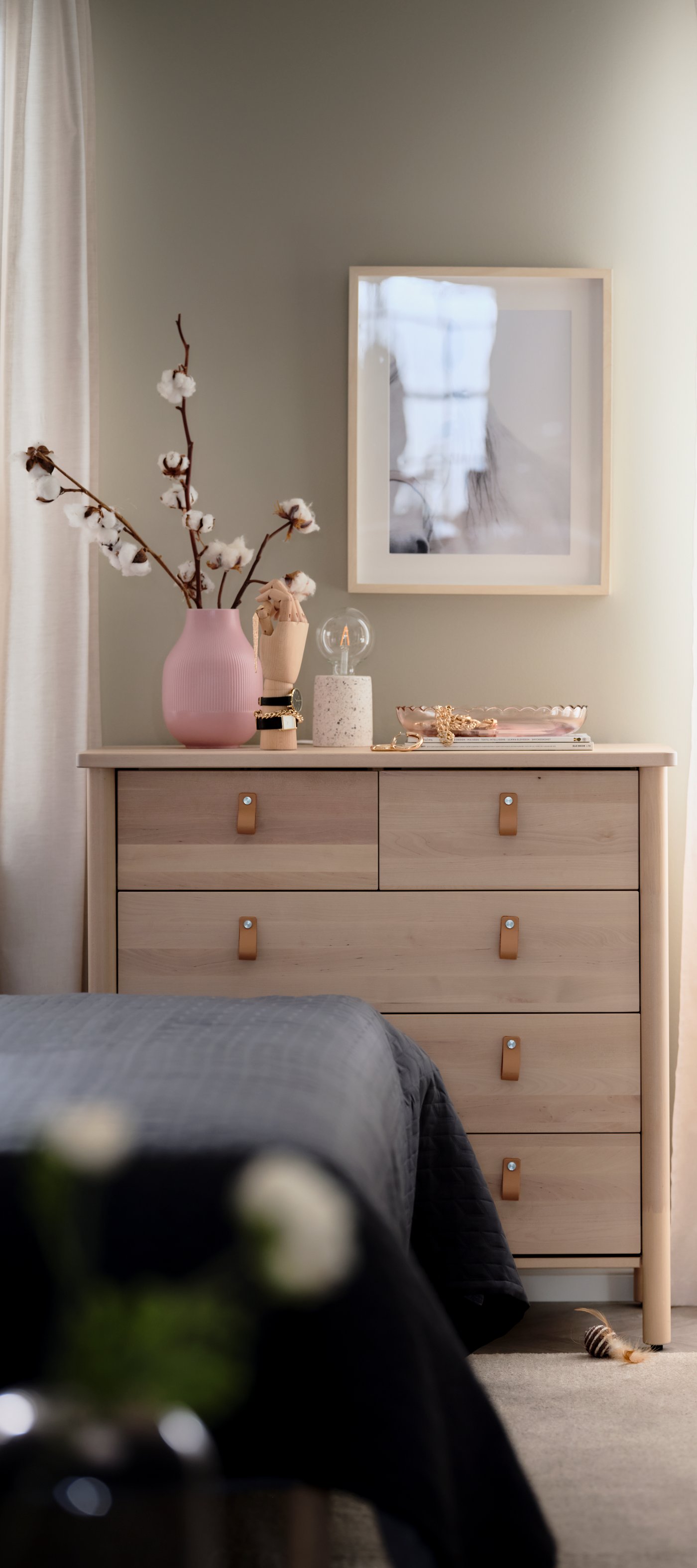 Ljus träbyrå med fyra lådor och läderhandtag. Ovanpå byrån står en rosa vas med bomullsgrenar, en liten dekorativ burk och andra små prydnadsföremål. Ovanför hänger en inramad tavla och till vänster syns en vit gardin och i förgrunden skymtar kanten av en säng med mörkt bäddset.