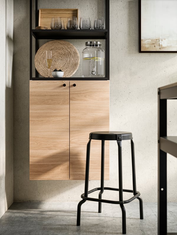 A RÅSKOG bar stool in black and an ENHET wall frame with shelves in anthracite holding diverse glasses, jars and other items.