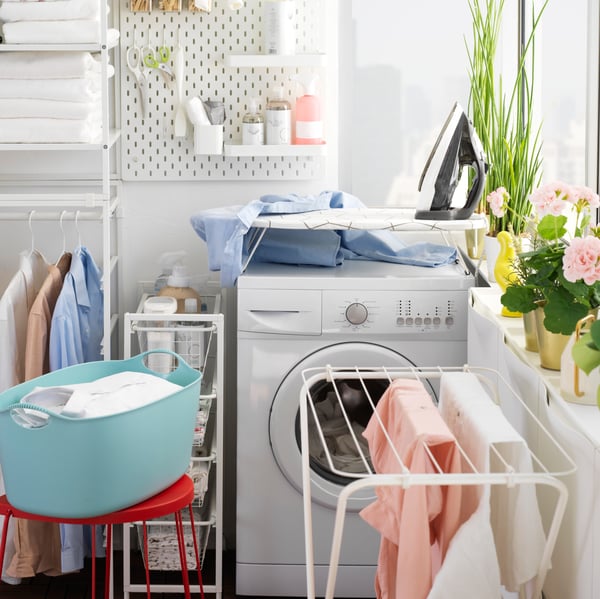 Clothes hang on a white JÄLL drying rack in front of a washing machine in a laundry room.