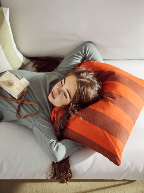 A girl lying on a sofa rests her head on the orange and brown-striped HILDAMARIA cushion cover.