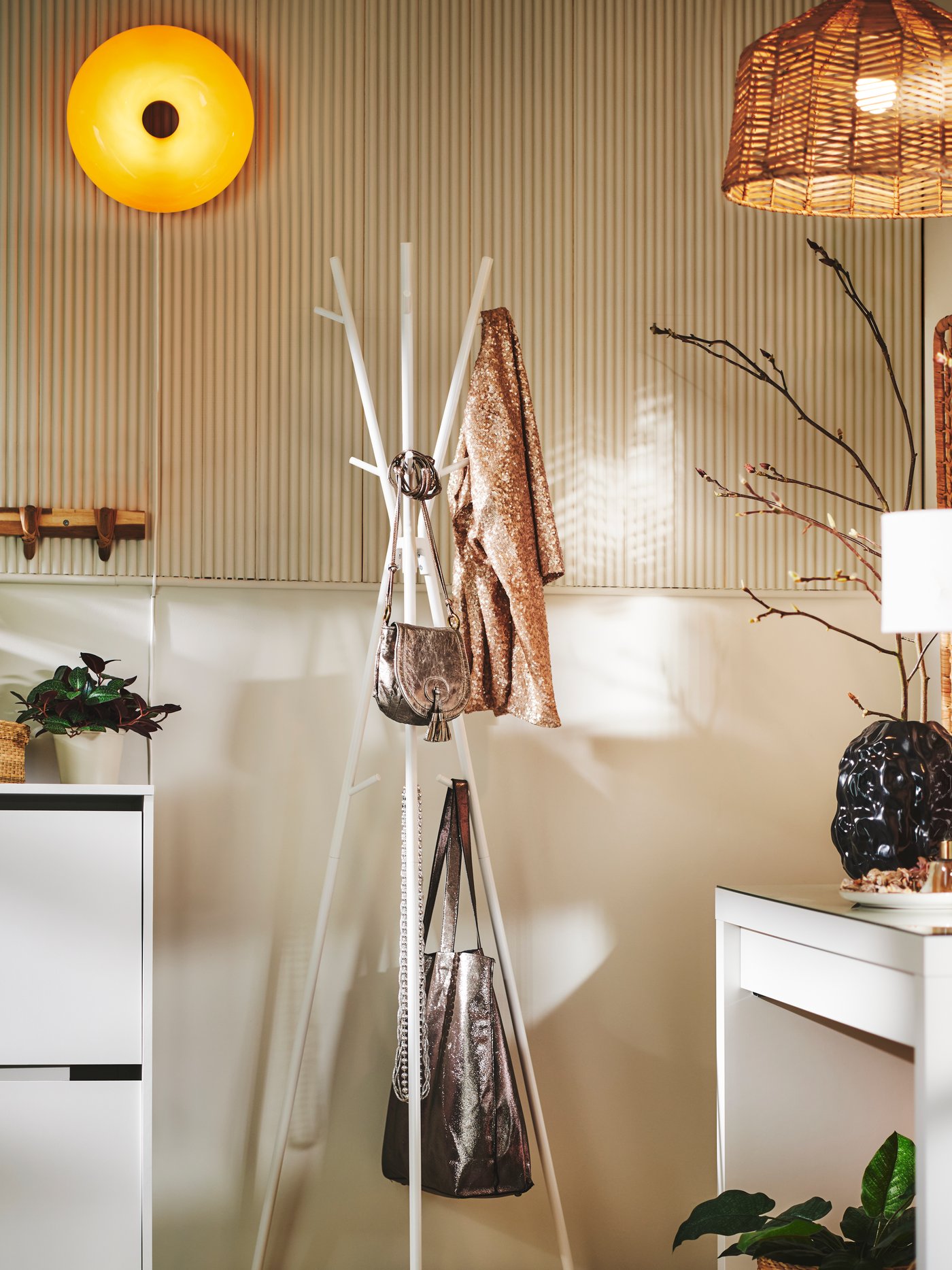 Ett vitt EKRAR hatt- och klädställ bredvid ett BISSA skoskåp håller olika saker, med en rund orange VARMBLIXT vägglampa ovanför.