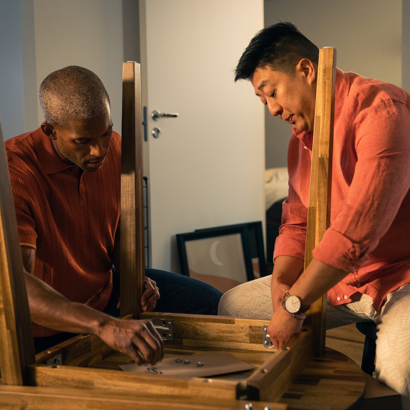 Two men screw together an Ikea table together while sitting on their knees wearing red shirts.