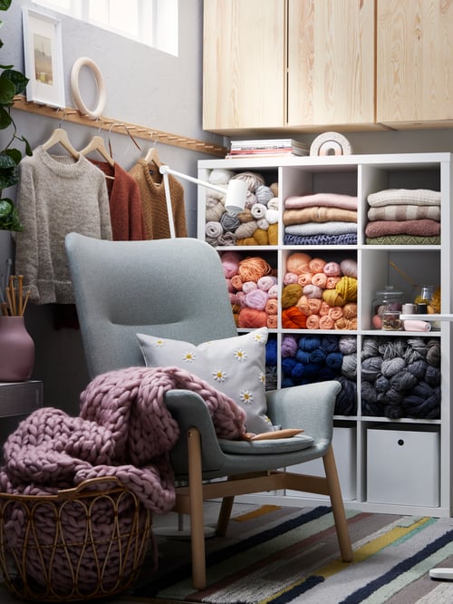 A light green VEDBO armchair with sweaters on hangers behind, a KALLAX shelving unit with yarn, plus pine cabinets above.