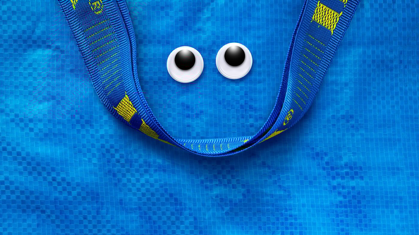 Eine blaue FRAKTA Tasche, bei der die Henkel als Mund dienen und Augen aufgeklebt sind. 