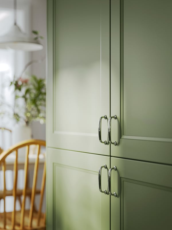 Façades AXSTAD gris-vert avec des poignées argentées, à côté d’une chaise en bois et d’une plante en arrière-plan.